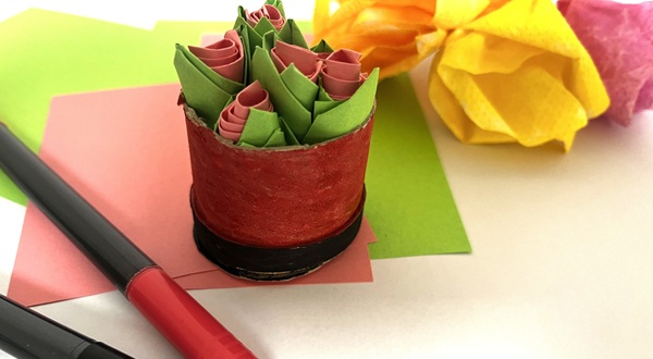 Handgjord minibukett av färgat papper i röd cylinder, omgiven av pysselmaterial och pennor.