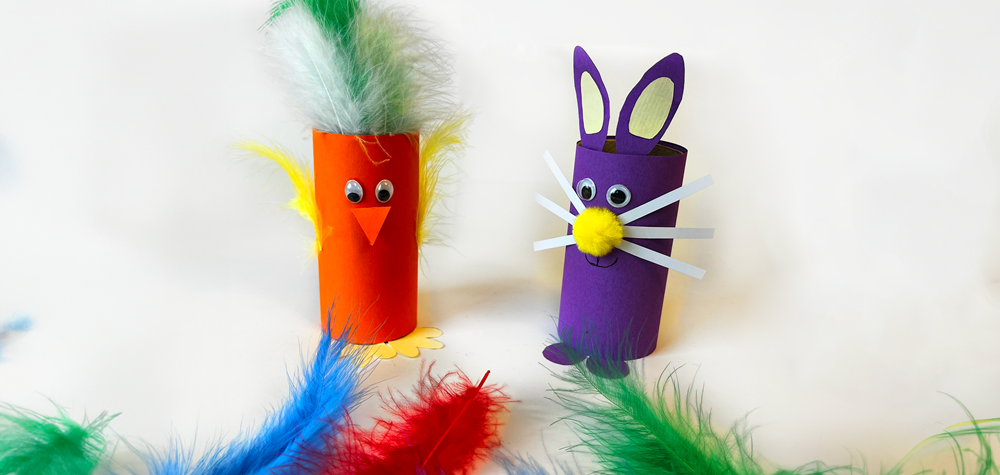 Två färgglada pysselfigurer gjorda av toarullar, en orange fågel med fjädrar och en lila kanin med morrhår, omgivna av färgade fjädrar
