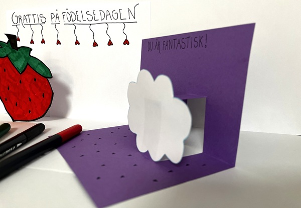 Handgjort 3D‑kort i lila med pop‑up‑moln och födelsedagshälsning, dekorerat med ritade detaljer och pennor.