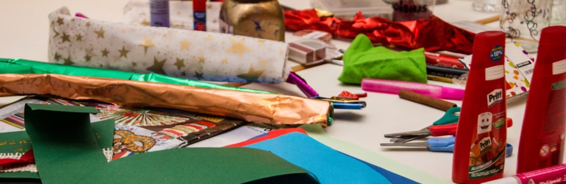 Julpysselmaterial på ett bord, färgat papper, presentpapper med stjärnor, lim, saxar, glitter och dekorationsband i rött och guld.