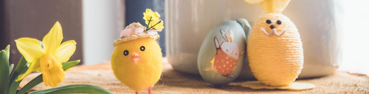 Påskpyssel med dekorationer, gul kyckling med hatt och blomma, gul kanin av garn och ett målat påskägg med kaninmotiv, placerade på träbord bredvid gula påskliljor.