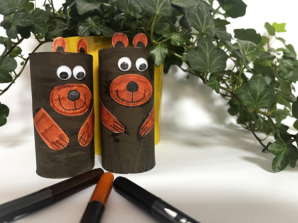 Två handgjorda björnar av målade toarullar med rörliga ögon, placerade framför gröna växter och omgivna av färgpennor.