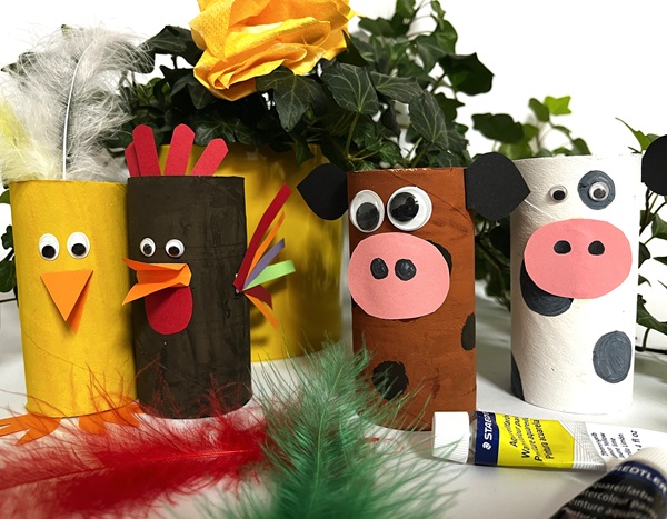 Fyra djurfigurer gjorda av målade toarullar: en gul kyckling med fjädrar, en brun tupp med färgglada vingar, en brun ko med rosa mule och en vit ko med svarta fläckar. Fjädrar och lim ligger framför, gröna växter och en gul blomma i bakgrunden.