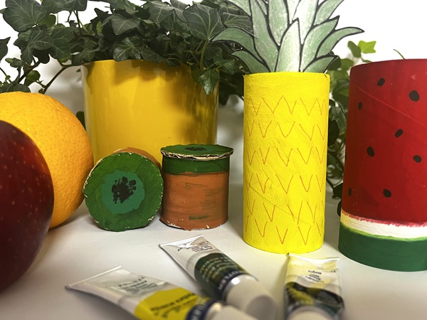 Färgglada fruktfigurer gjorda av målade toarullar: en gul ananas med gröna blad, en röd vattenmelon med svarta prickar och två små gröna och bruna frukter. Framför ligger tuber med färg, och i bakgrunden syns gröna växter samt en gul kruka.