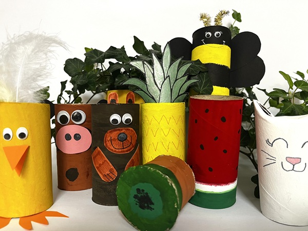 Flera färgglada figurer gjorda av målade toarullar, inklusive djur och frukter: en gul kyckling, en brun ko, en brun björn, en gul ananas, en röd vattenmelon, en svart-gul bi och en vit katt. Gröna växter syns i bakgrunden.
