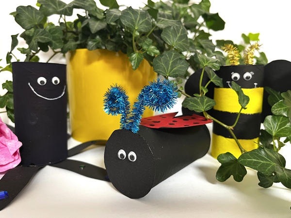 Tre insektsfigurer gjorda av målade toarullar: en svart figur med vitt leende, en liggande svart nyckelpiga med blå antenner och röda vingar med svarta prickar, samt en gul-svart bi med svarta vingar och guldfärgade antenner. Gröna växter och en gul kruka i bakgrunden.