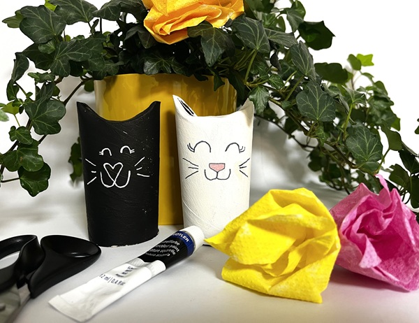 Två kattfigurer gjorda av målade toarullar, en svart och en vit, med ritade ansikten och spetsiga öron. Framför ligger en sax, ett limstift och färgglada pappersblommor, med gröna växter och en gul kruka i bakgrunden.